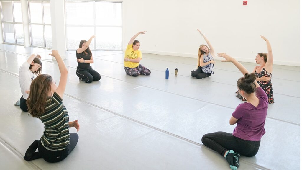 A few woman are sitting on the floor in a circle stretching their arm above their head to the opposite side of their arm stretching their side.