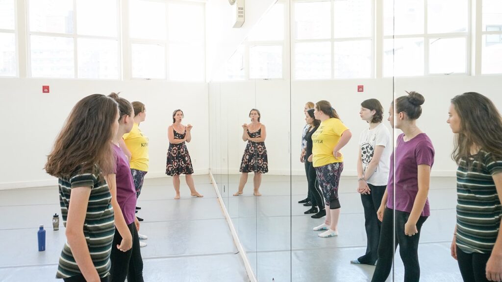 A few girls are standing in front of a mirror in a line looking at the instructor at the end.