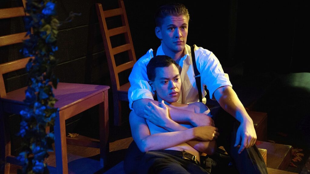 A man sitting on the floor in a white shirt and black suspenders holding another man who is laid in front of him with his head on his chest.