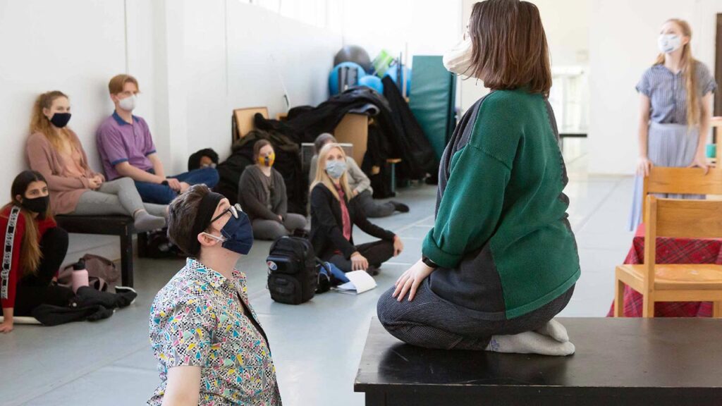 A woman kneels on a desk wearing a mask with a woman kneeled in front of her with a. mask on.