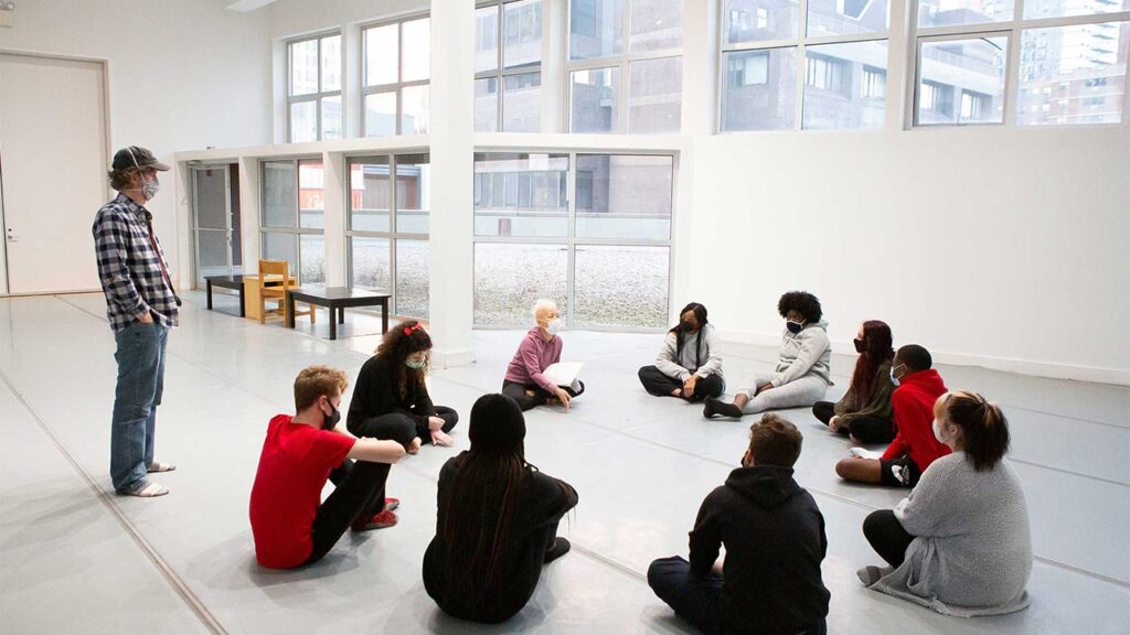 A group of people sit on the floor in a circle wearing masks to cover their nose and mouth. A man stands with his hands in his pockets outside of the circle.