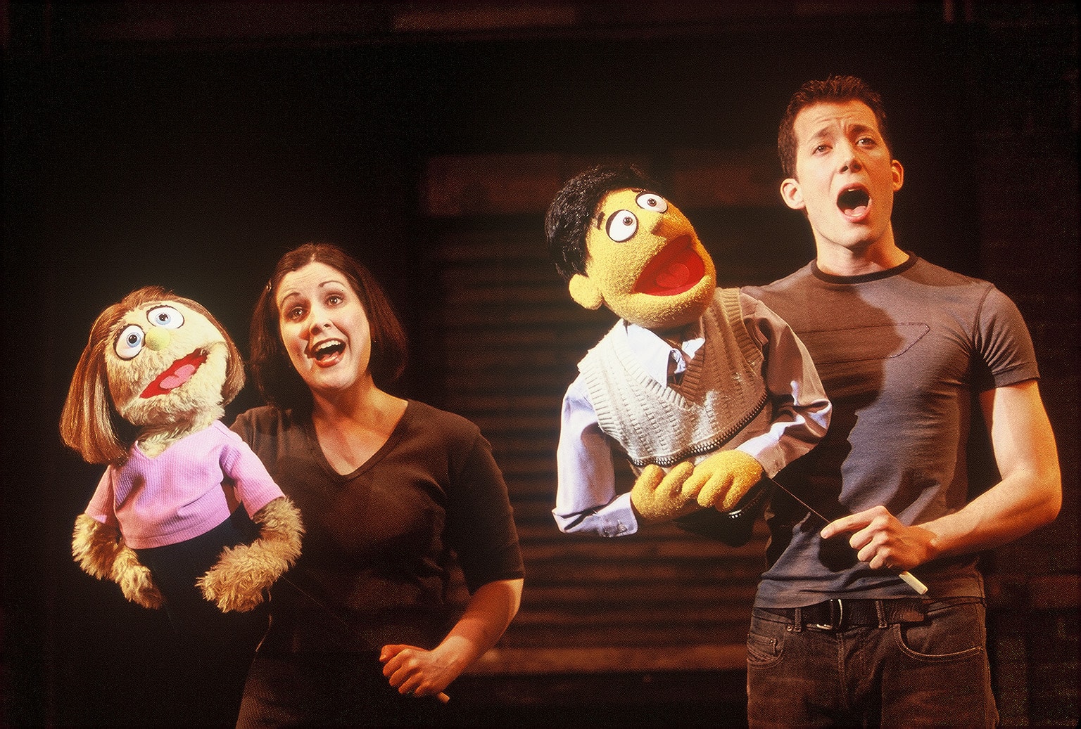 A woman and a man are standing on stage with each having a woman and a man puppet in their hands. All of are singing.