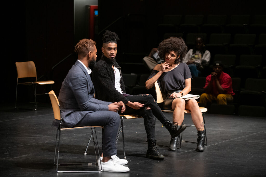 Three people sit on stage in chairs in a row. One man is talking while the other man and woman are looking at him. The woman has a pen and note book on her lap.