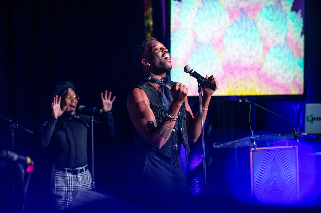 A large T.V screen in the background with colors of yellow, blue, pink, and white as a man stands with both hands in a fist singing.